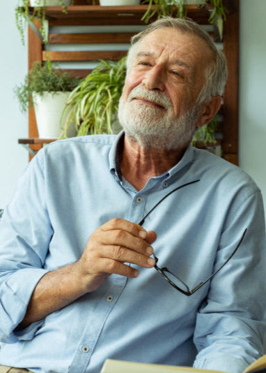 Man with a beard holding glasses, sitting indoors with plants in the background