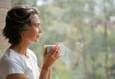 Woman holding a mug, looking out a window with raindrops on the glass