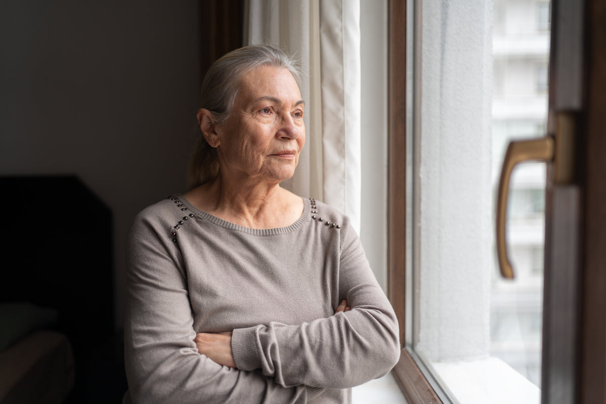 Senior woman standing by a window with arms crossed, looking out.